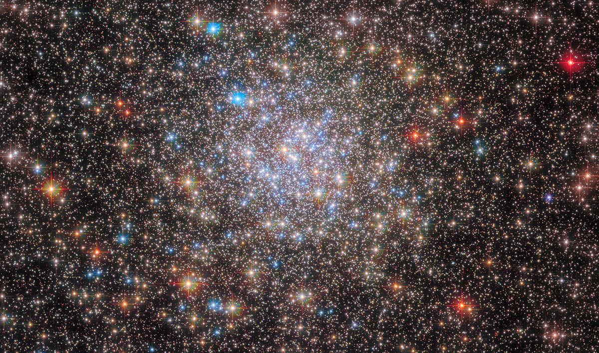 Crowded image of stars on black background. Stars of varying brightnesses and widths populate this image, with the most crowded cluster of bright-white stars centered around the middle of the image. Other stars are orange, blue, and red. The stars resemble bright pinpricks on a black canvas.