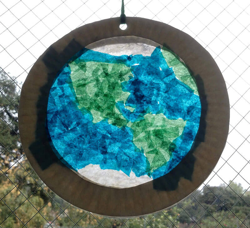 a photo of the Earth stained glass activity final product - paper glued to a white paper plate handing on a fence with the sky in the background.