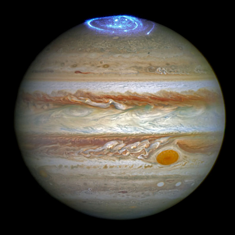 Auroras on the north pole of Jupiter. Full-disc view of colorful, banded clouds and red storm on Jupiter against a black background. The banded, swirling clouds look wispy, as if stripes of wet paint were painted and gently swirled. The aurorae on the top of the planet look like neon swirling fireworks.