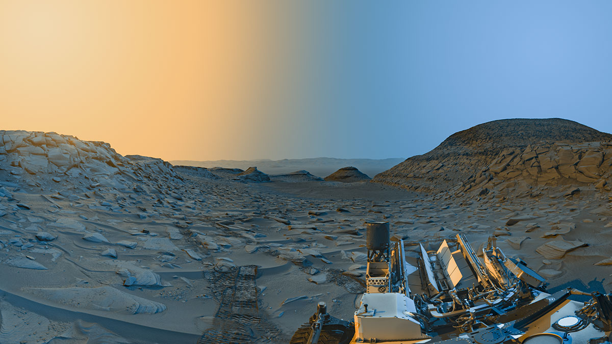 Colorized photo from NASA’s Mars Curiosity rover shows a wide view of a rocky valley on Mars. The rear half of the rover is visible in the lower right, with its wheel tracks on the surface receding into the distance. This stylized view adds color to a pair of black-and-white panoramas taken by the rover in the morning and afternoon. Blue and yellow tones simulate the lighting of the morning and afternoon, respectively.