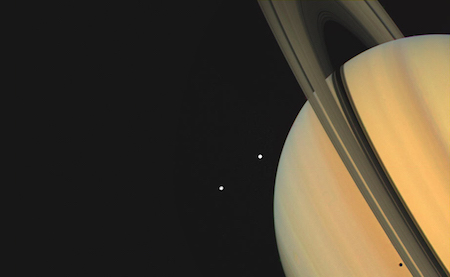 A photo of Saturn with its rings at an angle pointing upwards. Next to Saturn are two white dots, which are moons.