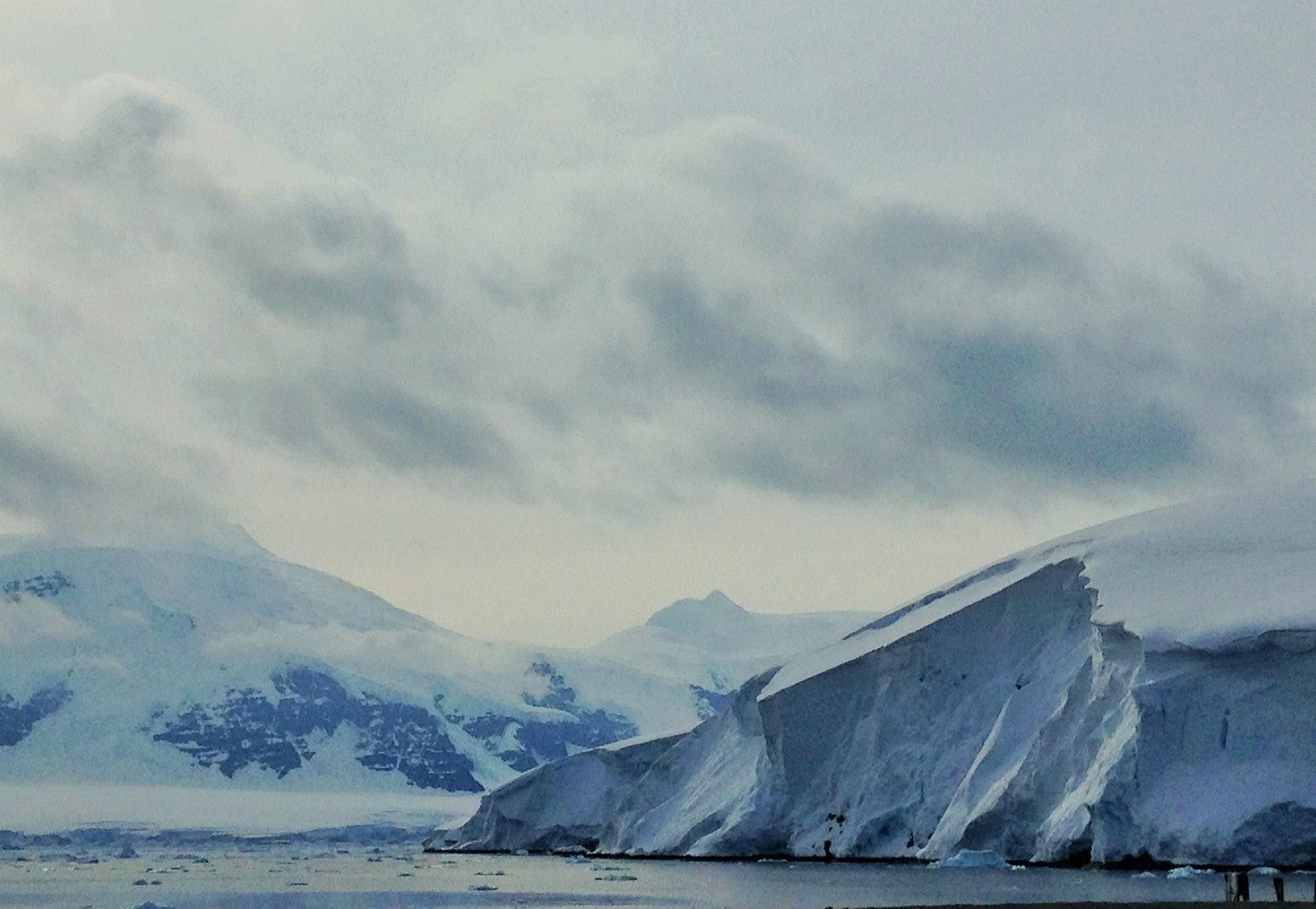 Antarctica’s terrain on a partly cloudy day. Icy, snow-covered mountains rise above the sea, while a huge snowy ice shelf appears closer.