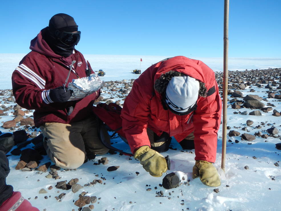 Members of the Antarctic Search for Meteorites (ANSMET) program collect a carbonaceous chondrite meteorite from a glacial moraine at the base of Mt. Ward, Antarctica. The two members are wearing red coats, as well as hats, mittens, and goggles. The member on the right is on their hands and knees, using a tool to investigate a meteorite rock. The member on the left is holding aluminum foil and another tool and observes the other team member.