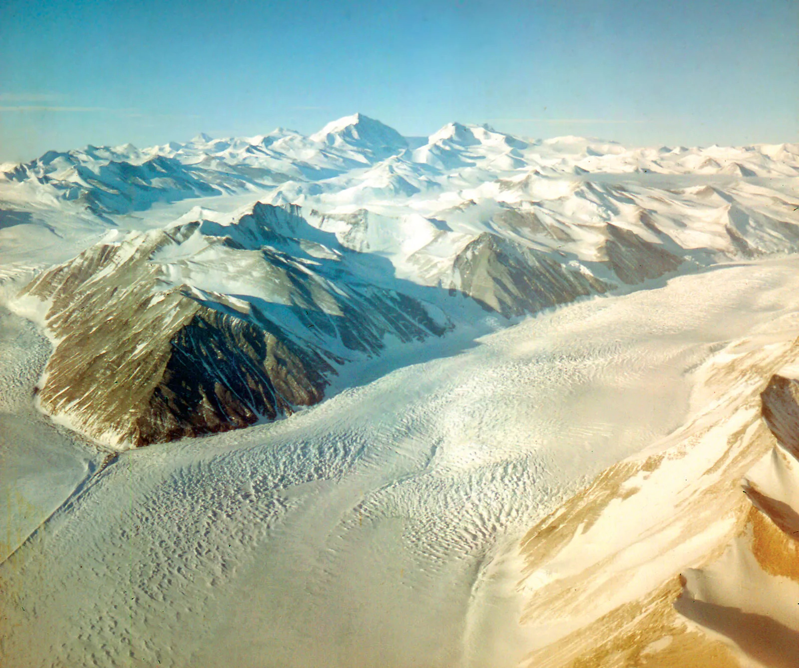 Beardmore Glacier in central Antarctica. The mountainous terrain forms a valley of snow and ice. Most of the mountaintops are covered in snow, but some are exposed and rocky terrain can be seen.