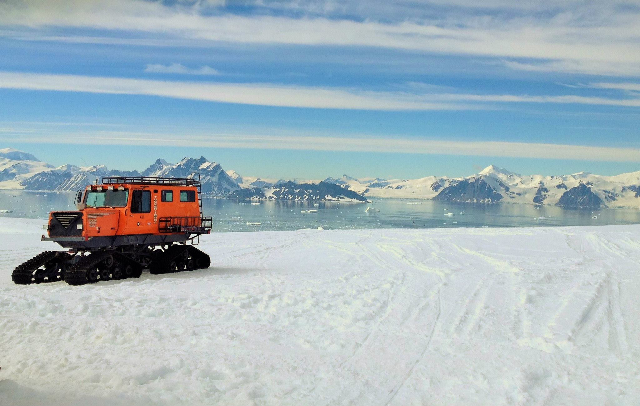 Antarctica’s bright white landscape. An orange snowmobile vehicle sits atop an icy white surface in the foreground. Wispy clouds line the sky above the mountainous and icy landscape.
