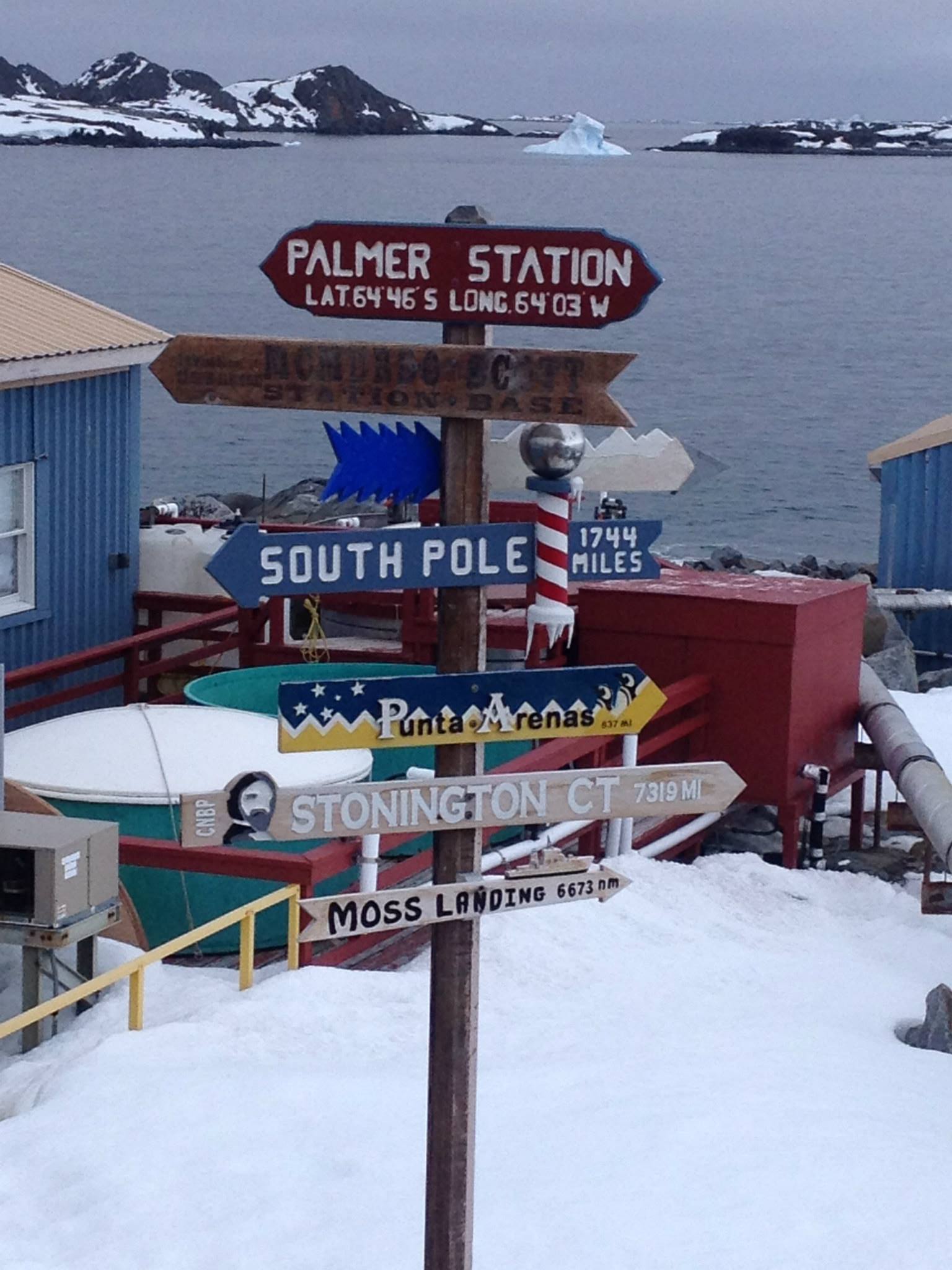 Signs in Antarctica point to different locations on Earth and show how many miles away that location is. For example, the South Pole points to the left of the image and is 1744 miles away. The ground is covered in snow, and icy terrain rises from the sea in the background.