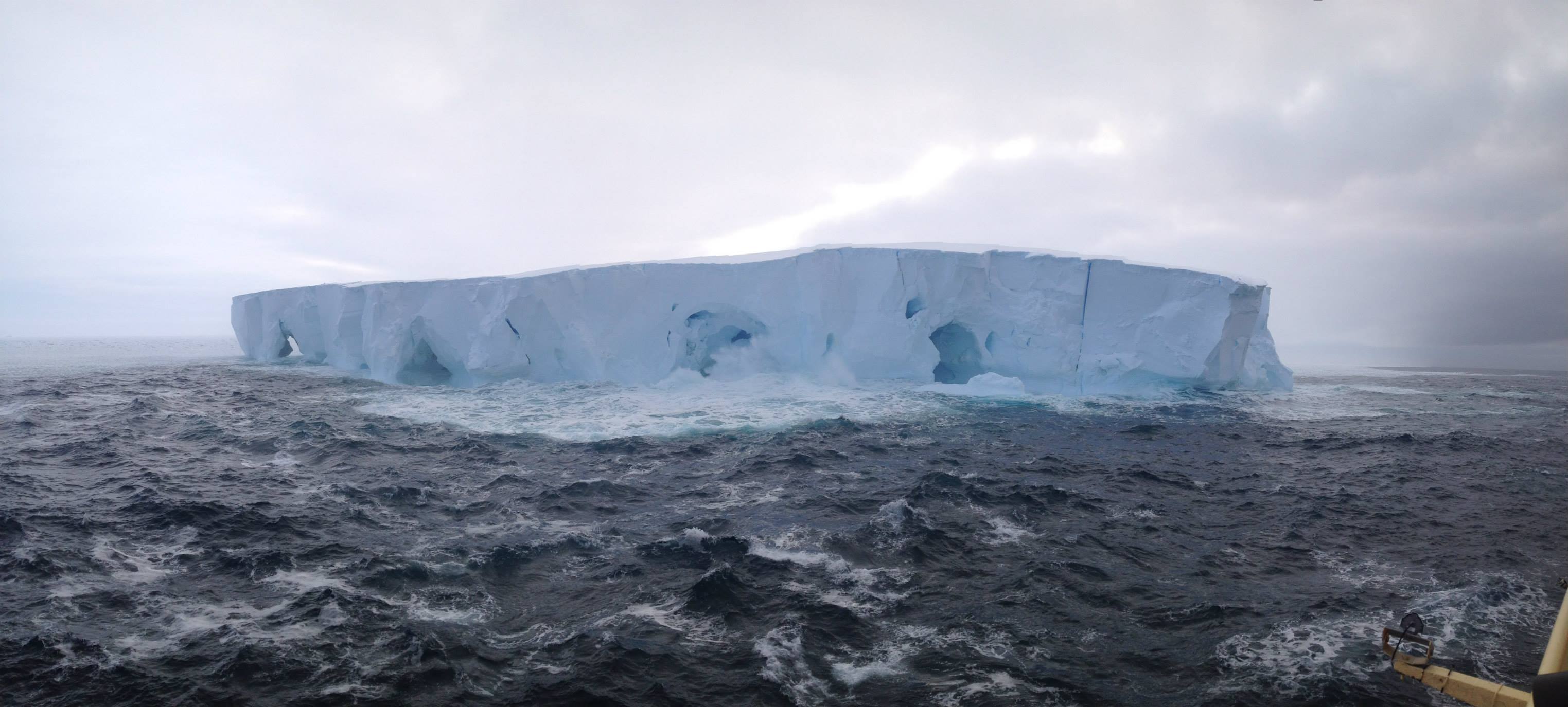 A bright white glacier in the dark blue choppy waters of Antarctica against a cloudy sky.