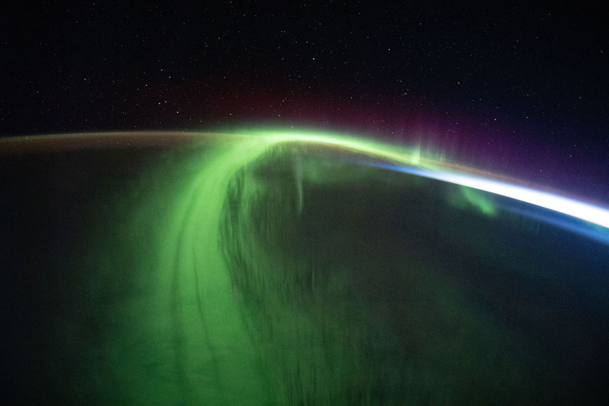 Green streaks of auroras arch over the dark Earth. A glint of white sunlight marks the curve of Earth in the horizon.