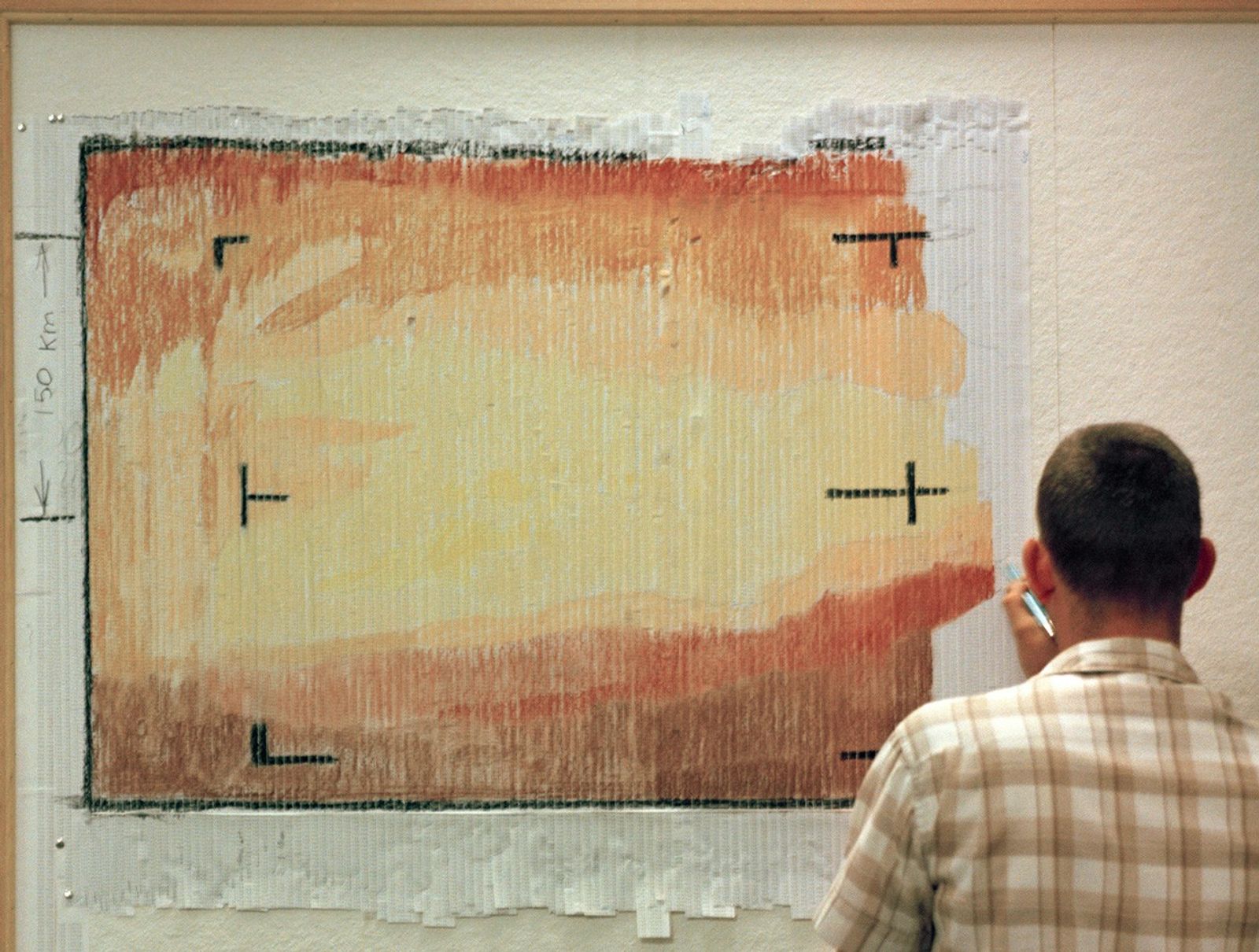 A man with short hair and a yellow-and-white plaid shirt stands with his back to the camera. In front of him, taped to the wall are long strips of paper side by side, each about 3 feet long. The strips have been colored different shades of yellow, orange and brown to create a picture with a dark band of brown along the bottom, and a wide yellow band running left to right, flanked by narrower bands in shades or orange.