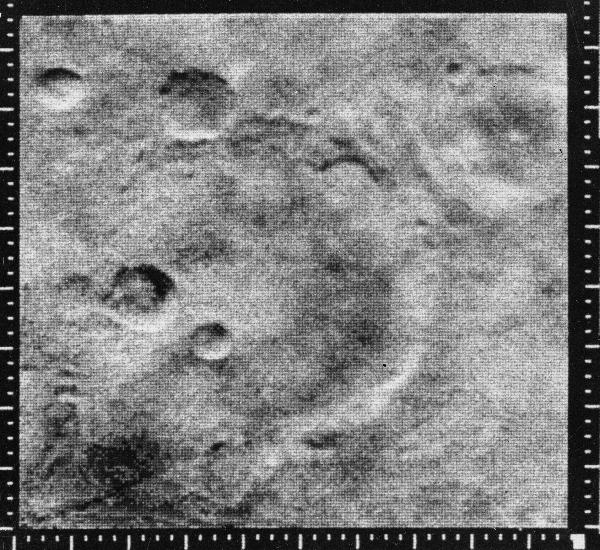 A grayscale photo of the Martian surface shows barren ground with several craters, large and small; the largest one, in the center, has two smaller craters inside it. Measurement markings line the outside edges of the frame, at the bottom and left and right sides.