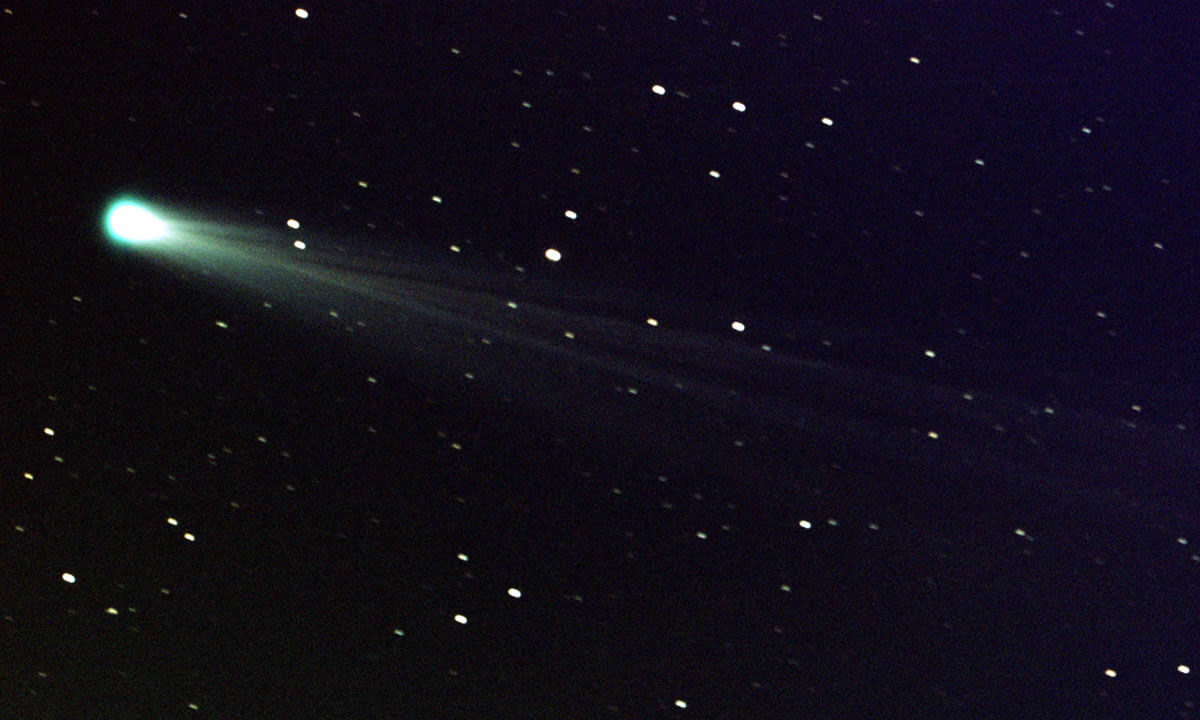 A black background is dotted with tiny points of white light, which are stars. At left is a large, bright, hazy object with streaks of bright dust streaming away from it toward the right.