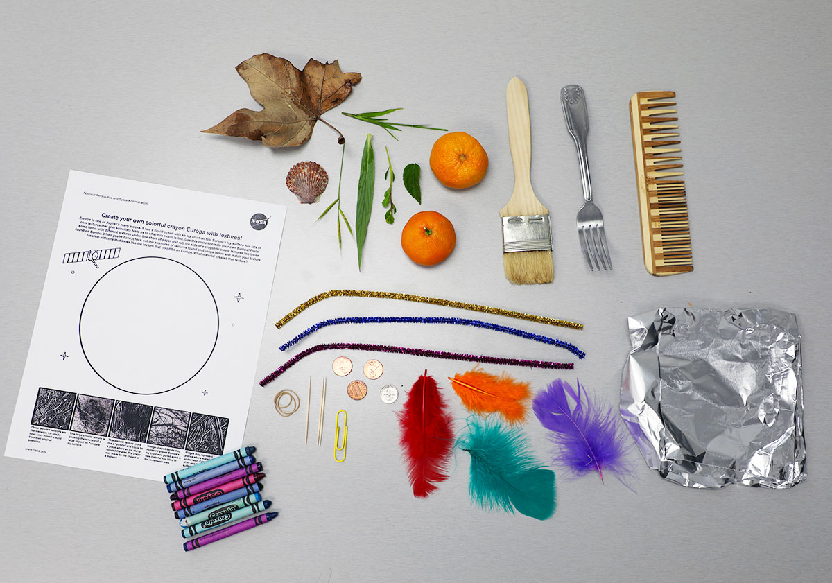 Materials needed for this activity laid out on a gray table. This image was taken from bird’s-eye view. The materials laid out on the table include two orange tangerines, a silver fork, four multi-colored feathers, a yellow paperclip, a small seashell, a paintbrush, a wooden comb, a small square of aluminum foil, a rubber band, blades of grass, pipe cleaners, toothpicks, three pennies and a dime, and the Europa activity sheet and eight crayons of various colors.