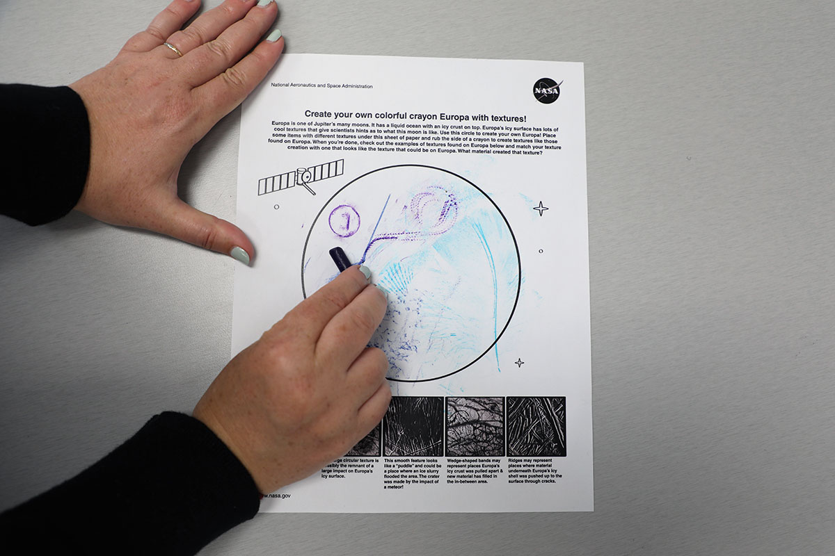 Hands rubbing a purple crayon against the Europa activity page.