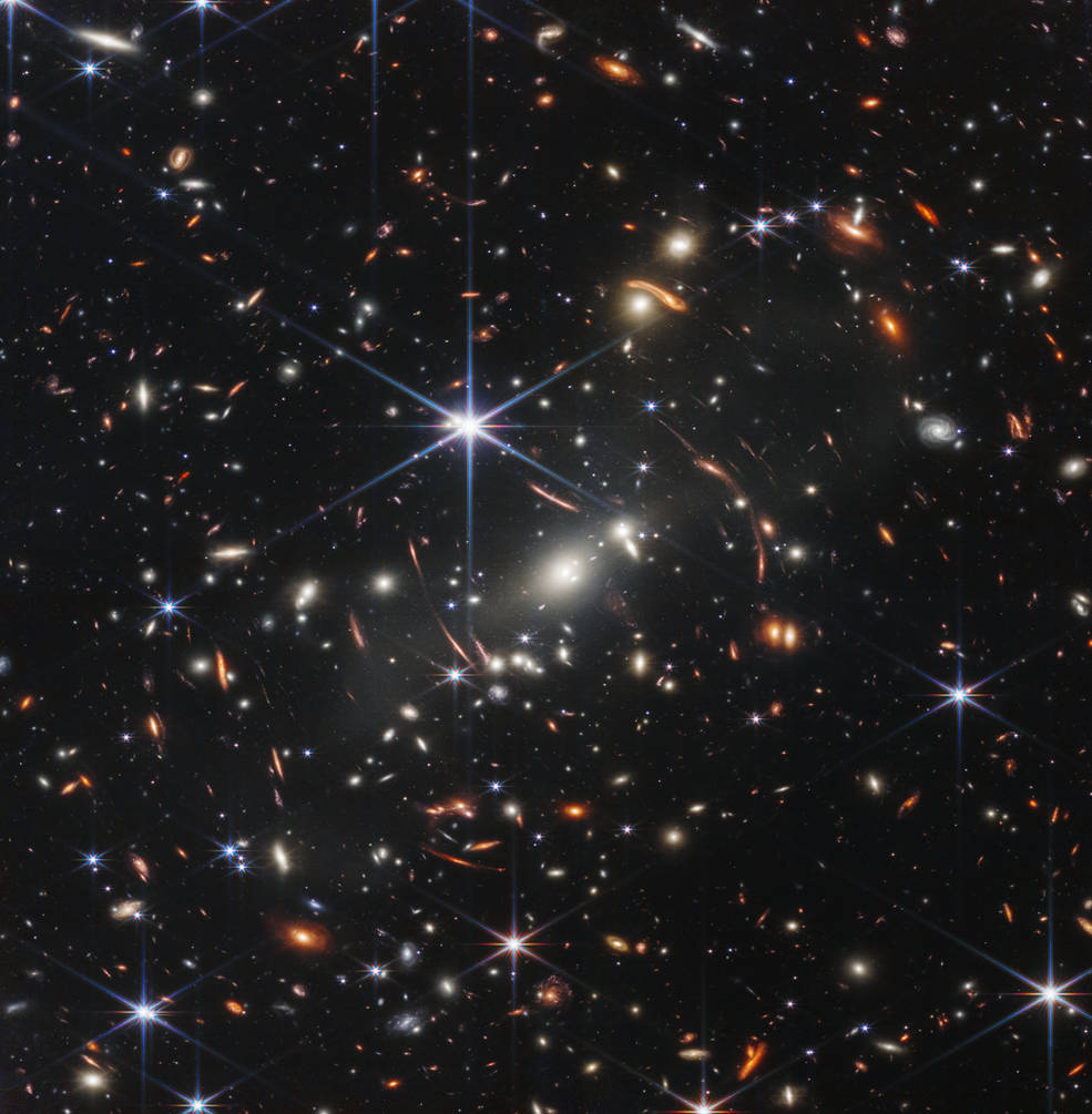 Distant galaxies appear as bright glowing spots in this Webb telescope image, with some smeared by gravitational lensing; foreground stars appear bright with six-pointed diffraction spikes, owing to the shape of Webb's mirrors.