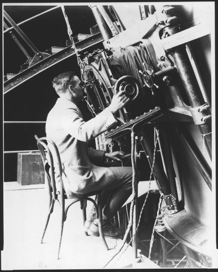 This black-and-white image shows Edwin Hubble sitting in a chair while looking through and operating a 100-inch reflecting telescope.