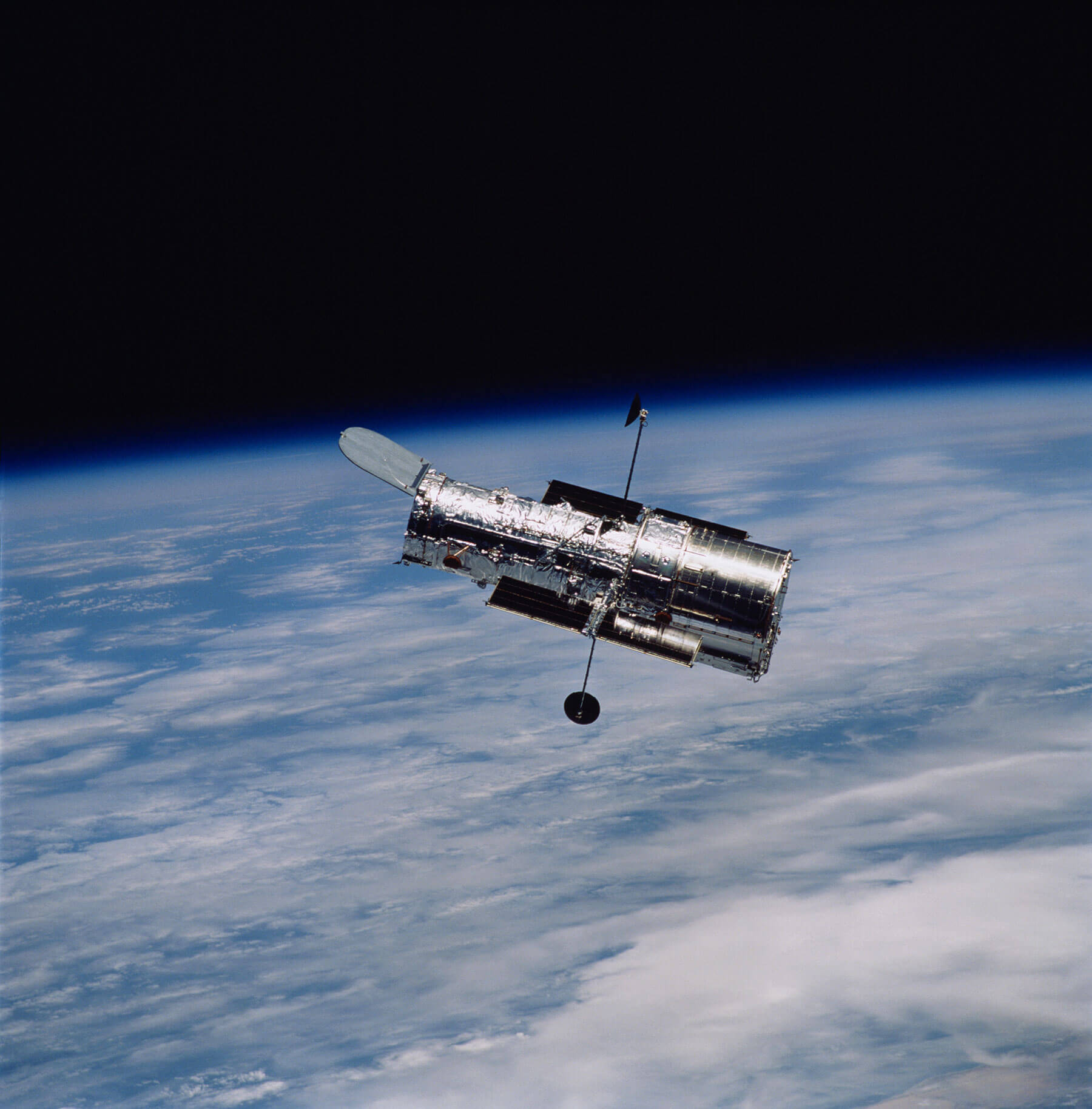 This is an image of the Hubble Space Telescope. A portion of Earth’s surface and horizon appear in the background. White wispy clouds can be seen intermittently covering Earth’s blue surface. The Hubble Space Telescope appears in the foreground. It is cylindrical in shape and has two antenna-like arms with a disc attached to each antenna’s end. The telescope itself is metallic gray and looks as if it is covered in aluminum foil.