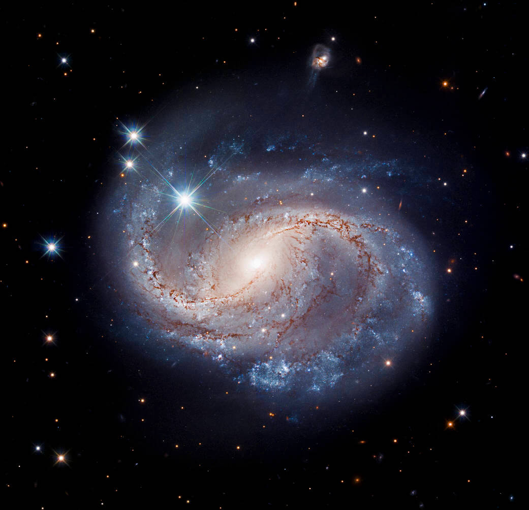 In the center of the image are blue and pinkish-white swirls of the barred spiral galaxy NGC 6956. Dark, reddish-brown dust lanes appear along the inner part of the spiral arms. The rest of this image consists of an inky black background dotted with white distant stars and galaxies.