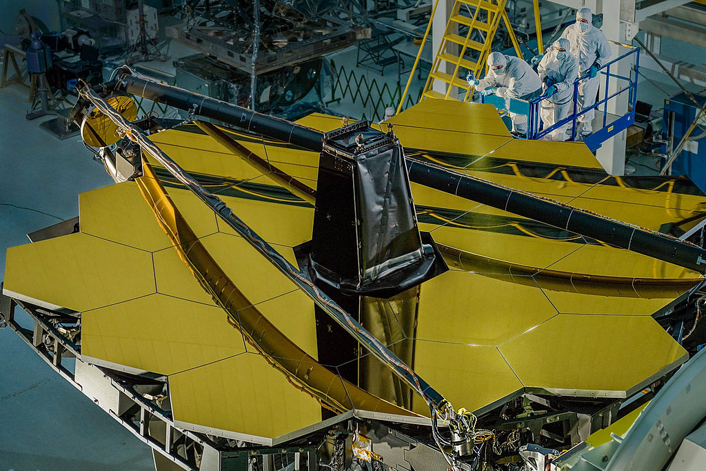The giant golden mirror of the James Webb Space Telescope made up of smaller golden hexagons. Three engineers in white clean room suits are standing on a platform to examine the mirror.