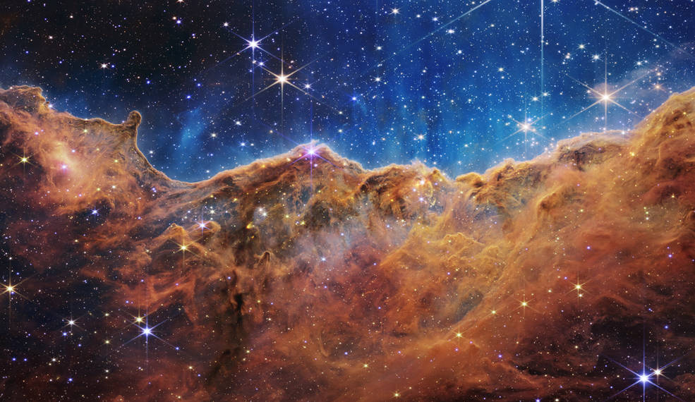 En esta imagen de Webb se muestra una región de formación de estrellas translúcida y ondulada en la Nebulosa de Carina, teñida de ámbar y azul; Se pueden ver estrellas en primer plano con picos de difracción, al igual que un moteado de puntos de luz de fondo a través de la nebulosa nublada.
