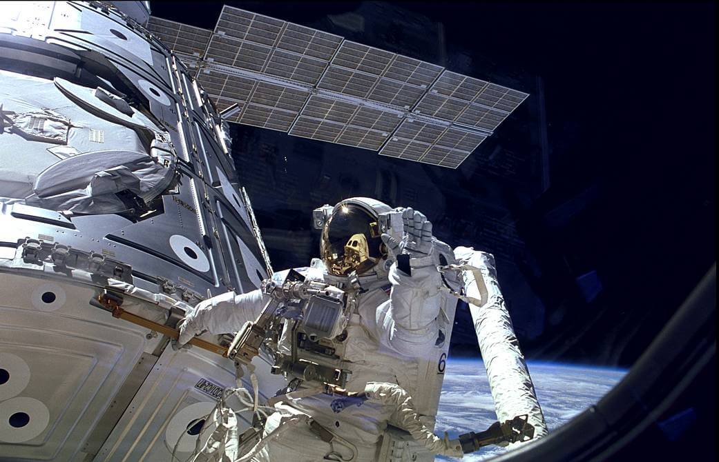 Astronaut James H. Newman waves during a spacewalk preparing for release of the first combined elements of the International Space Station. The astronaut wears a big white space suit and reflective helmet and waves at the camera in the foreground. The ISS’ solar panels and metallic body are behind him. Earth’s horizon is in the distance, and the rest of the background is deep, dark space.