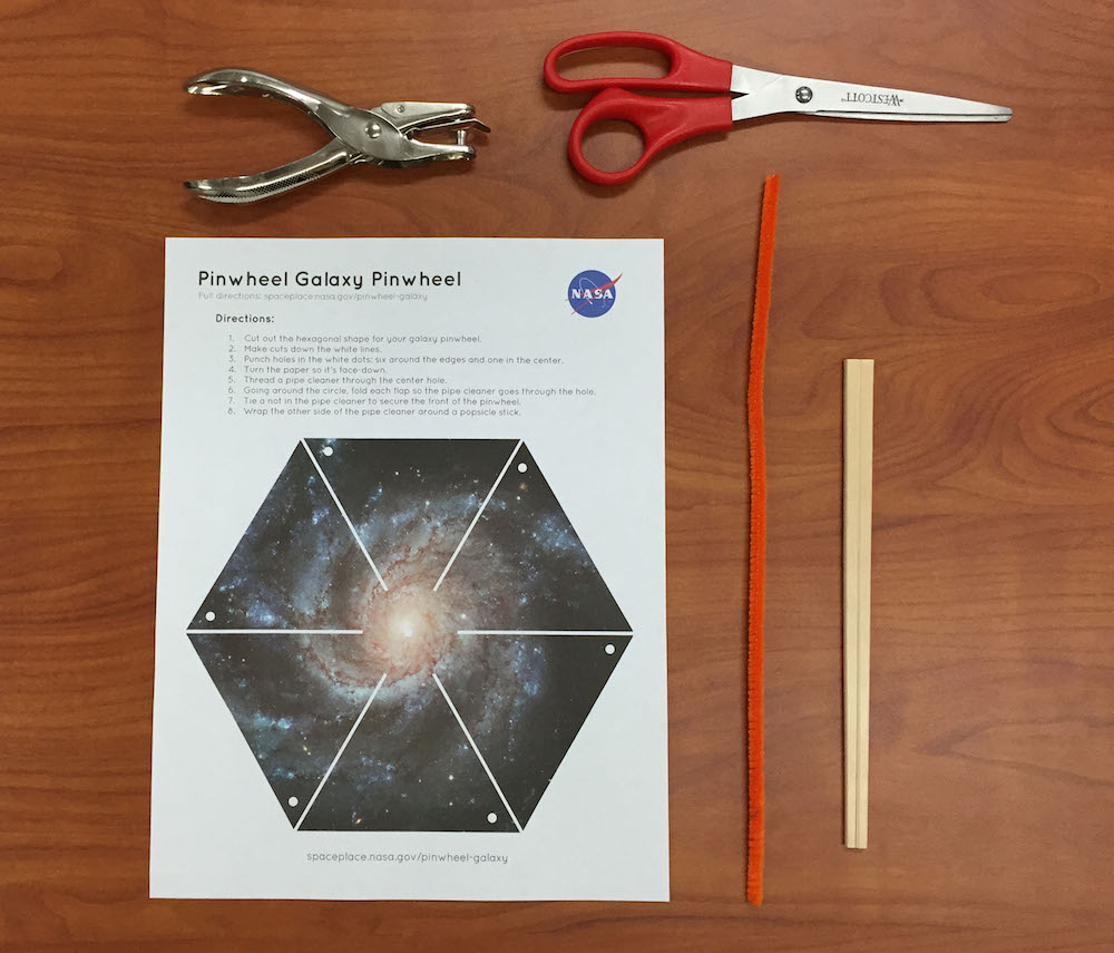 a photo of the supplies needed for this activity: the pinwheel galaxy printout, scissors, single hole puncher, a pipecleaner, and chopsticks.