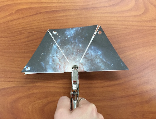 The hexagonal shape of the pinwheel galaxy printout folded in half with the hole puncher centered on it about to punch a hole.