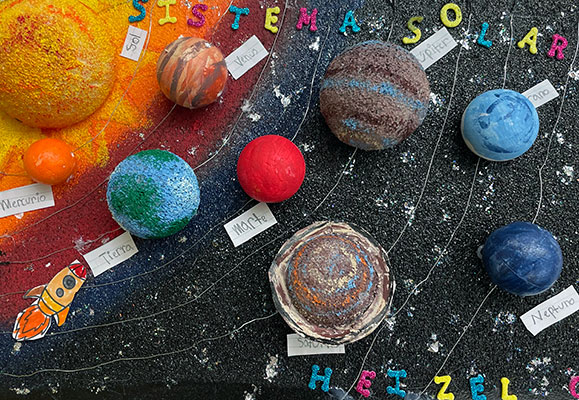Three-dimensional model of the solar system labeled in Spanish, Sistema Solar. The solar system is accurately represented in its order of planets. The planets appear to be made out of foam balls that have been cut in half, and painted to represent each planet’s respective colors. Each planet is labeled in Spanish. A cut-out of a rocket is glued on the background next to Earth.