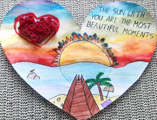 This valentine is in the shape of a heart. It shows a beach landscape during a sunset. Using watercolor paints, the artist recreated the colors of a sunset. Starting from the top of the valentine, the sunset sky colors include violet, blue, yellow, orange, and red. A yellow Sun with orange spiky rays sets behind a wavy light blue ocean. In the foreground, a brown pier extends from the edge of the valentine cutout to the ocean. A person is seen standing at the end of the pier, just before it ends. A sandy beach, complete with a purple towel, yellow umbrella, green palm tree with three coconuts, and some shells sits under the pier. On the left side of the ocean, a small brown bird sits atop a gray rock. Lastly, the valentine's text says, The sun with you are the most beautiful moments.