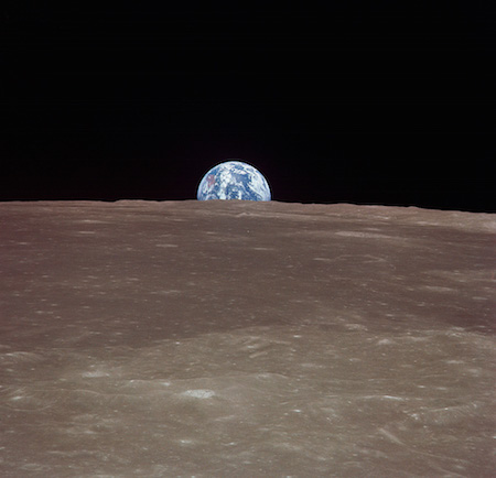 A photo of Earth in the background looking very small. The moon's surface is in the foreground, so Earth is rising over the moon.