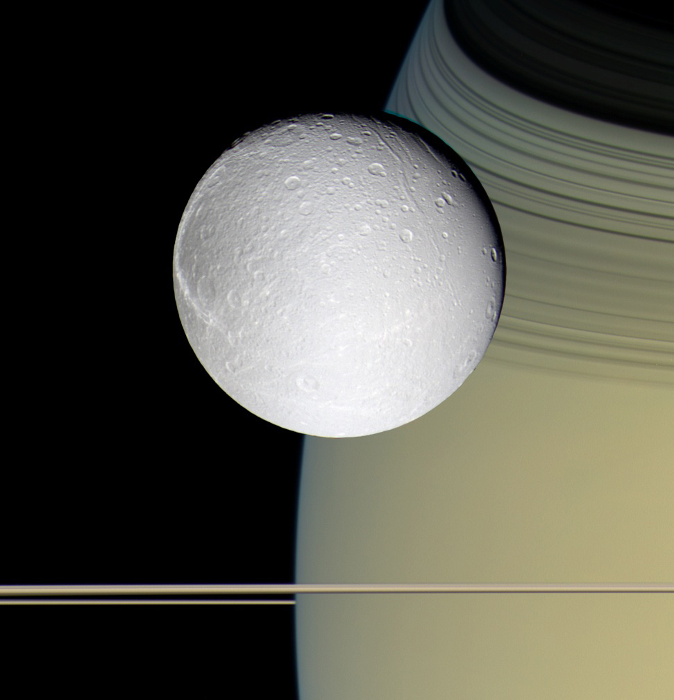 Una Dione blanca parece flotar en el espacio, con Saturno y sus anillos en el fondo.