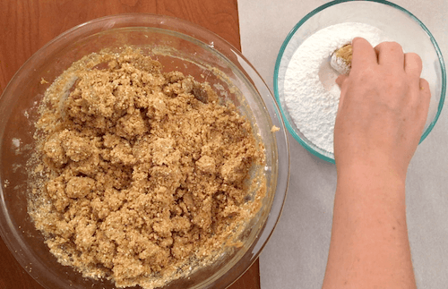 a person dips a cookie dough ball into a small bowl of powdered sugar.