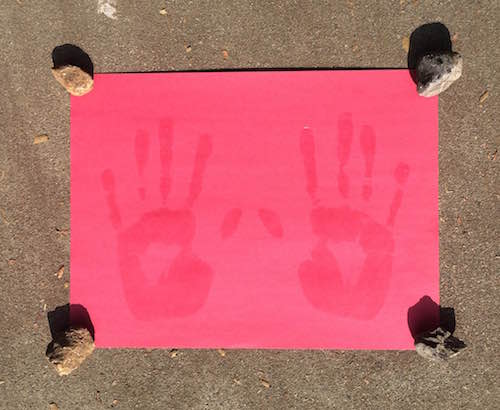 construction paper with wet sunscreen handprints placed in the sun with small rocks on the four corners of the paper