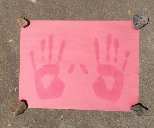 A piece of construction paper that is faded, except for two bold red handprints. There are four small rocks in each corner of the paper.
