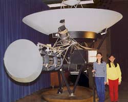 Foto de dos niñas de pie junto a modelo de tamaño real de la nave espacial Voyager, que es aproximadamente cuatro veces más alta que las niñas.